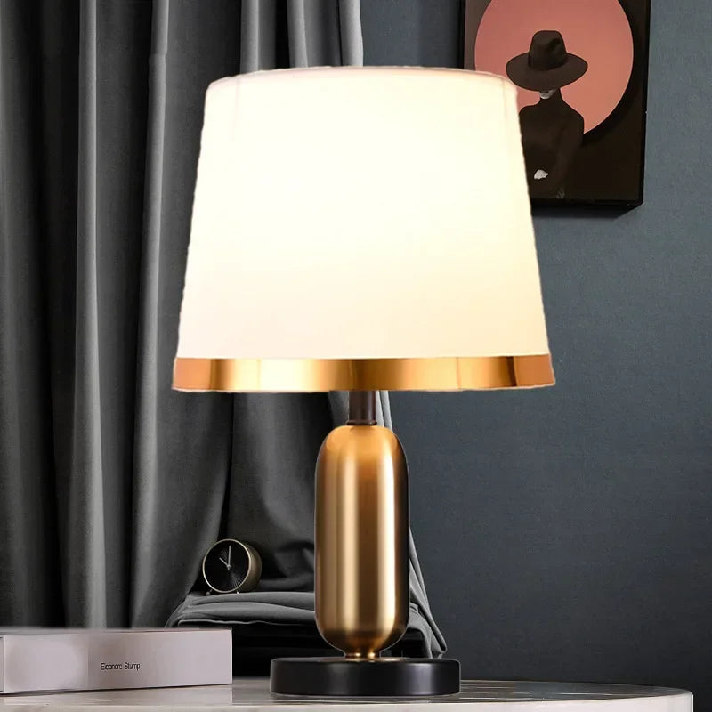 Table lamp with gold base and white shade on a surface against a dark curtain background